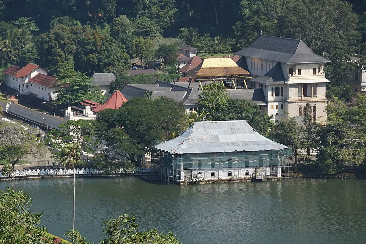 Kandy Lake in the foreground with the white-walled Temple of the Tooth complex and wooded hillside on the far shore, part of one building under scaffolding